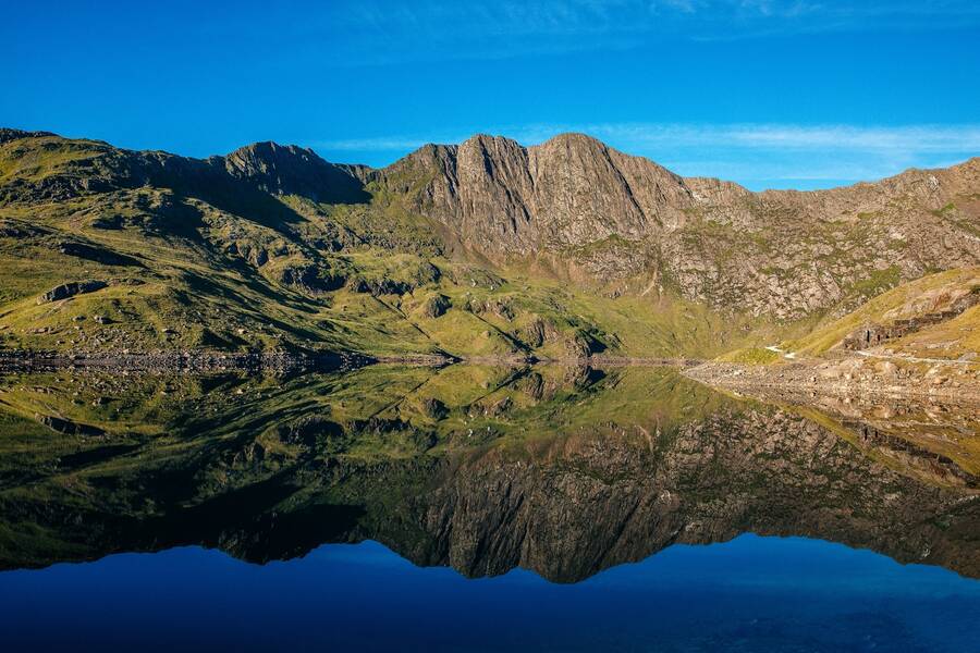 Still mountain lake reflecting rugged Snowdonia peaks and green slopes beneath a clear blue sky.