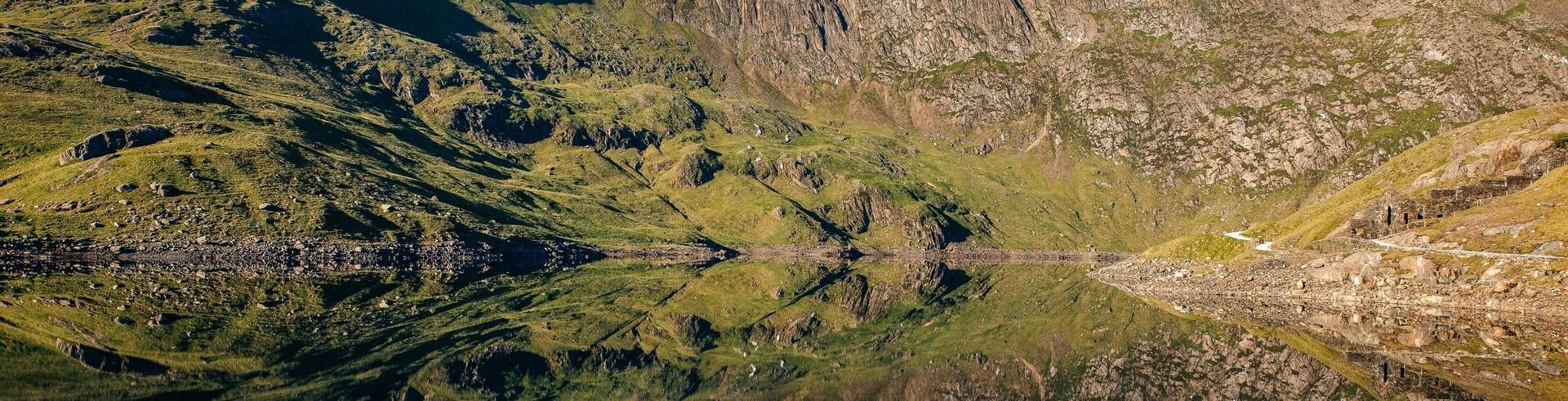 Still mountain lake reflecting rugged Snowdonia peaks and green slopes beneath a clear blue sky.