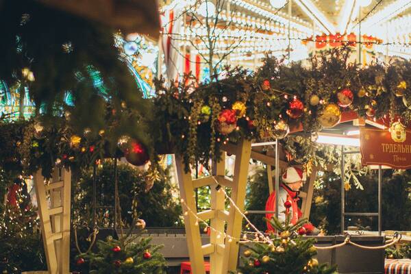 Traditional Christmas market stall decorated with lights, garlands and small fir trees, with a carousel glowing in the background at night.
