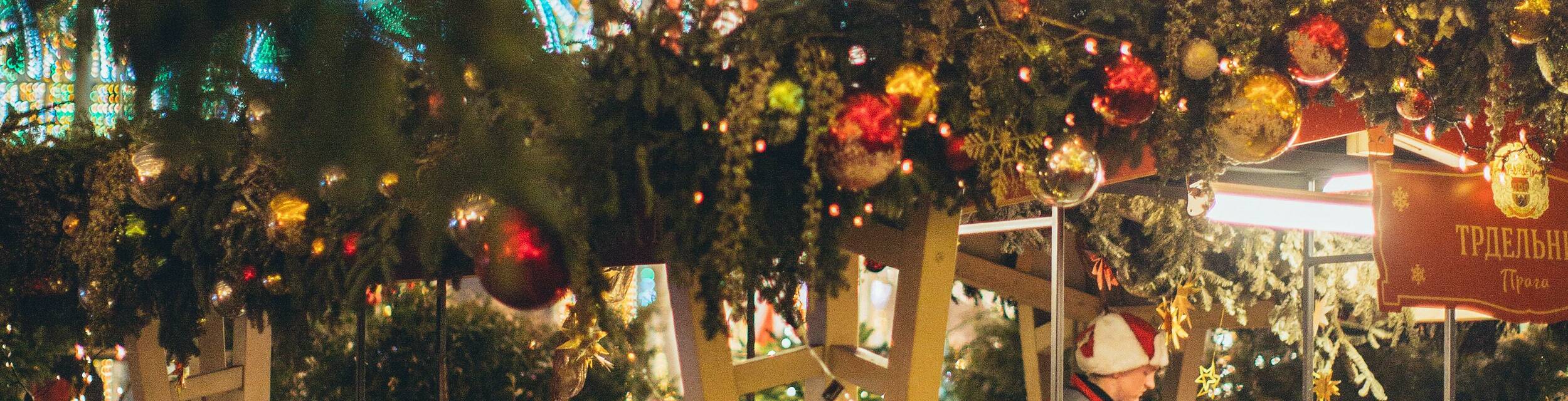 Traditional Christmas market stall decorated with lights, garlands and small fir trees, with a carousel glowing in the background at night.