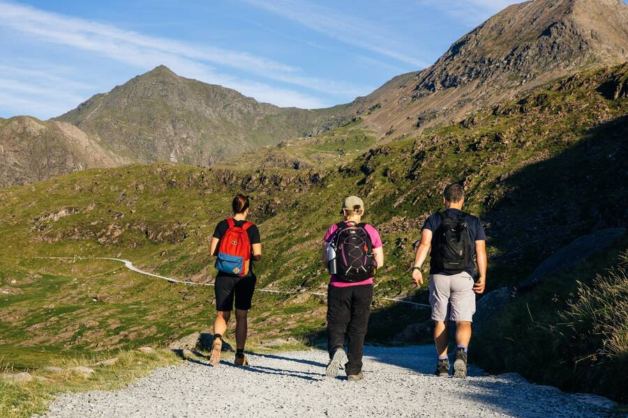 Three walkers with backpacks walking along a gravel mountain path with rugged peaks and valleys around Llanberis.