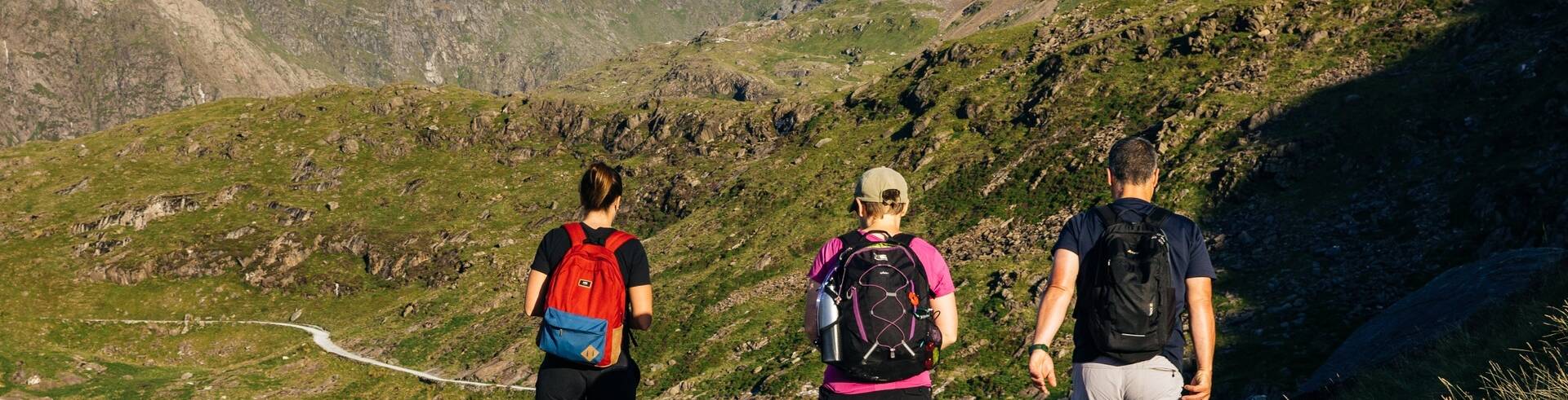 Three walkers with backpacks walking along a gravel mountain path with rugged peaks and valleys around Llanberis.