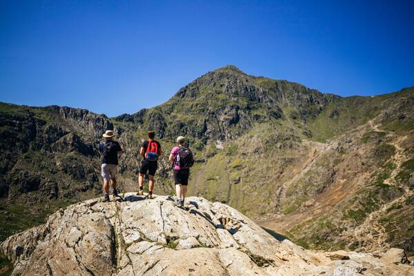 Three hikers standing on a rocky outcrop, looking towards the summit of Yr Wyddfa (Snowdon) under a clear blue sky.