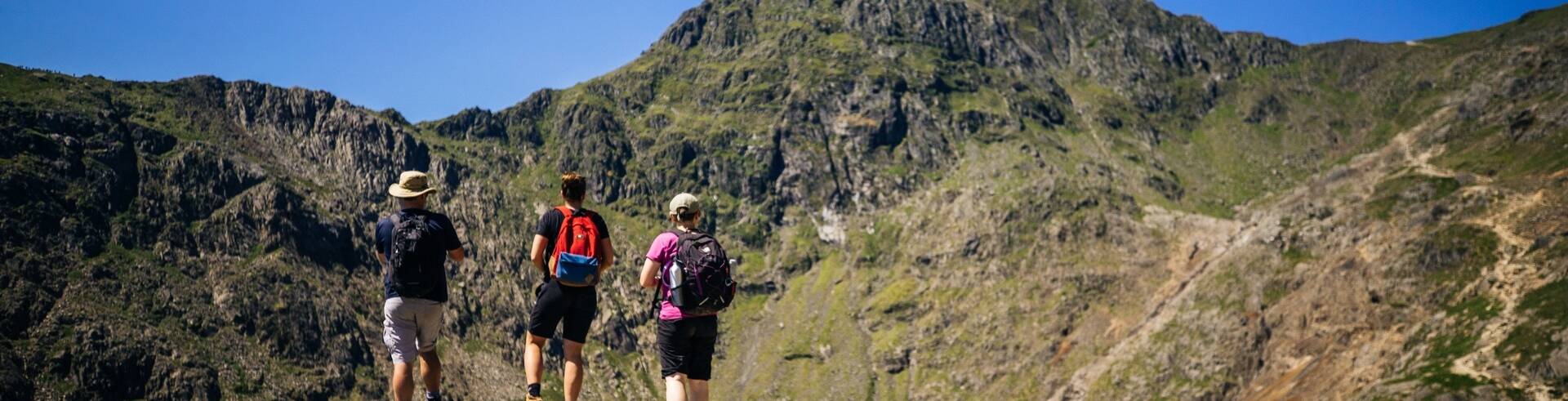 Three hikers standing on a rocky outcrop, looking towards the summit of Yr Wyddfa (Snowdon) under a clear blue sky.