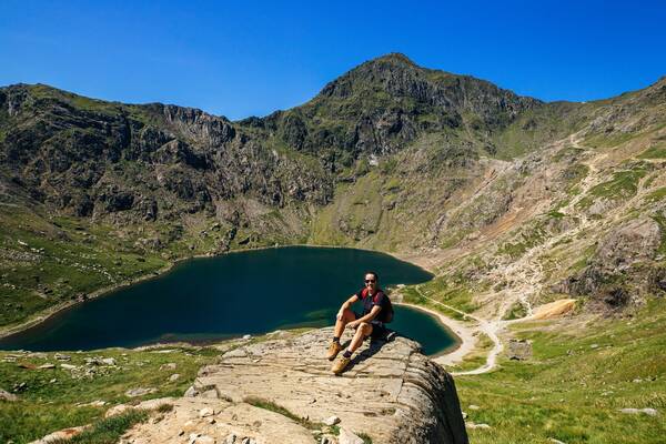 Hiker sitting on a rocky outcrop overlooking Llyn Llydaw with mountain scenery in Snowdonia National Park.
