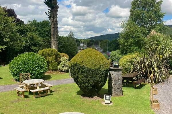 A well-kept hotel garden with wooden picnic tables, manicured shrubs and trees under a partly cloudy sky.