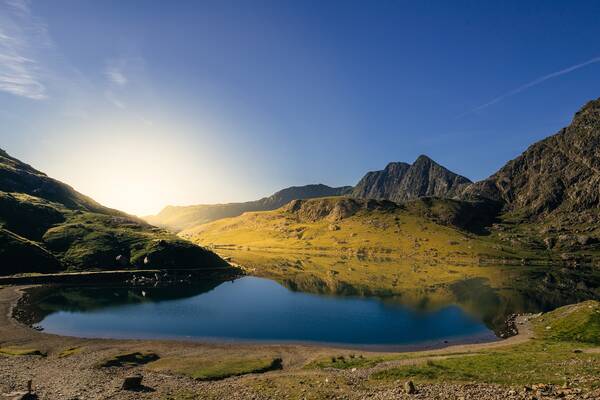 A calm mountain lake reflecting surrounding peaks and golden hills under a clear blue sky in Snowdonia.