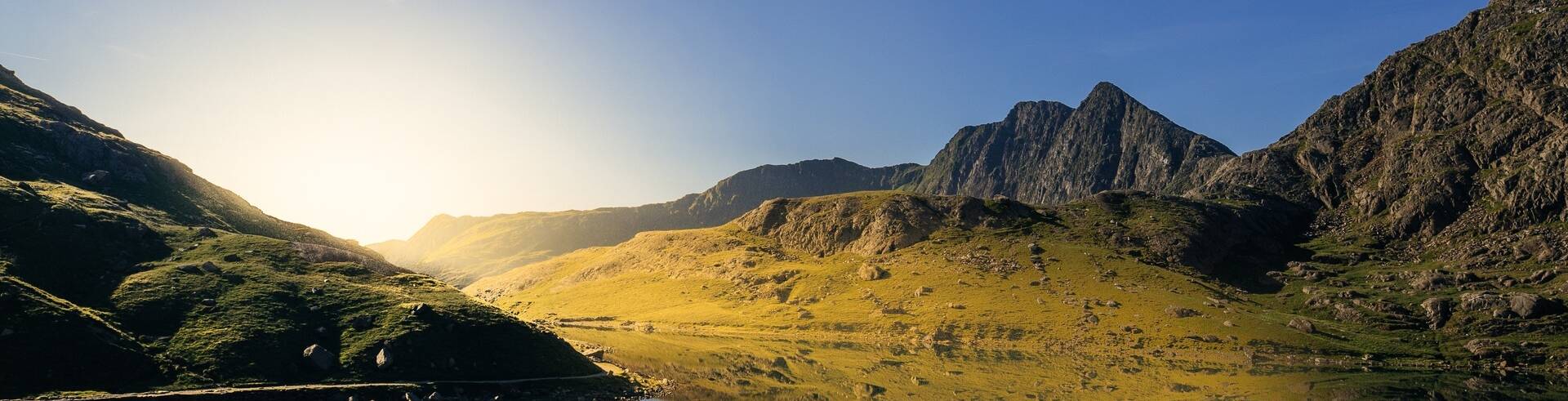 A calm mountain lake reflecting surrounding peaks and golden hills under a clear blue sky in Snowdonia.
