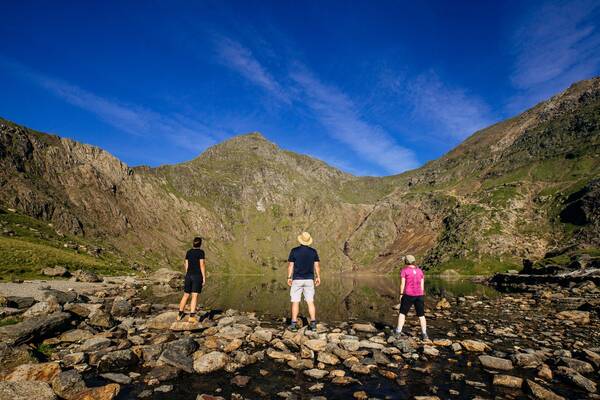 Three people standing on rocky ground beside a calm mountain lake, surrounded by steep peaks in Snowdonia.