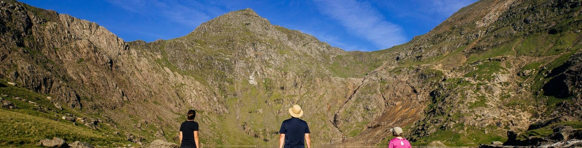 Three people standing on rocky ground beside a calm mountain lake, surrounded by steep peaks in Snowdonia.