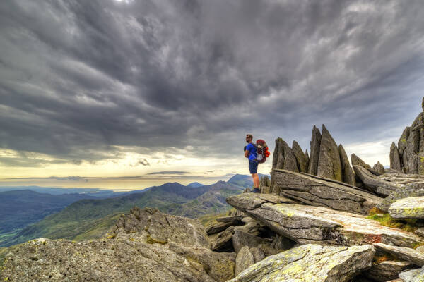 Hiker standing on jagged rocks near a mountain summit in Snowdonia under dramatic storm clouds.