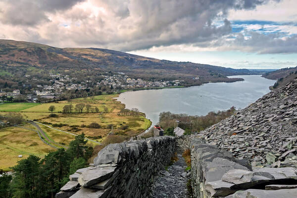 Slate path overlooking Llyn Padarn with the village of Llanberis, fields, and hills stretching into the distance under cloudy skies.