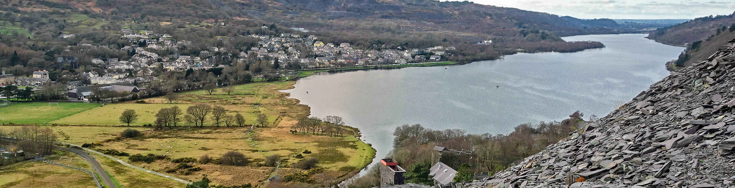 Stone slate path overlooking Llyn Padarn with the village of Llanberis, fields, and hills stretching into the distance under cloudy skies.