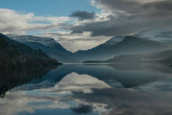 Snow-topped mountains surrounding Llyn Padarn near Llanberis are reflected perfectly in the still water, beneath a moody sky of clouds and soft light.