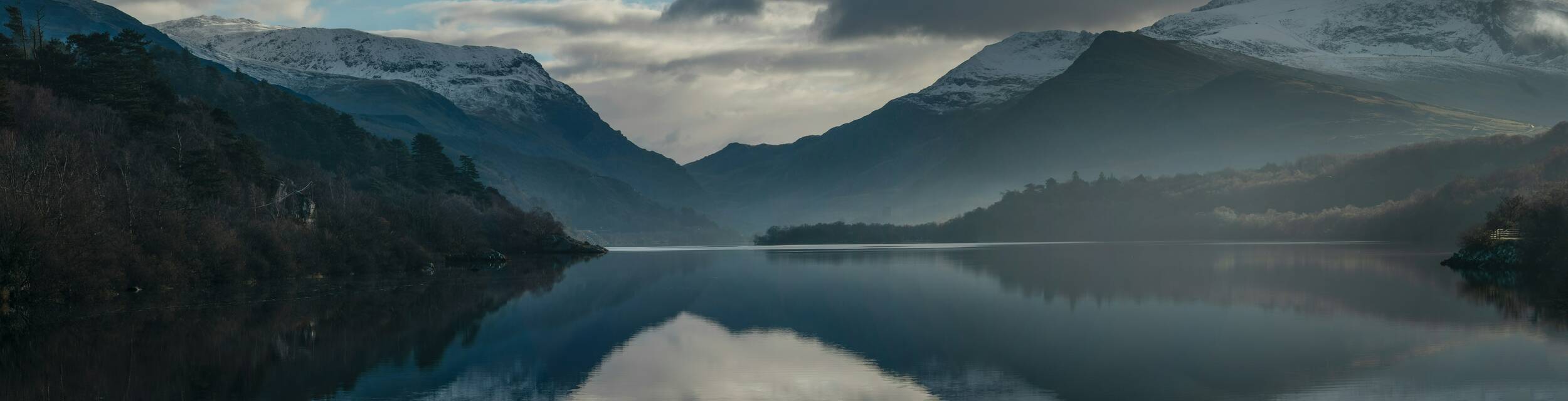 Snow-topped mountains surrounding Llyn Padarn near Llanberis are reflected perfectly in the still water, beneath a moody sky of clouds and soft light.