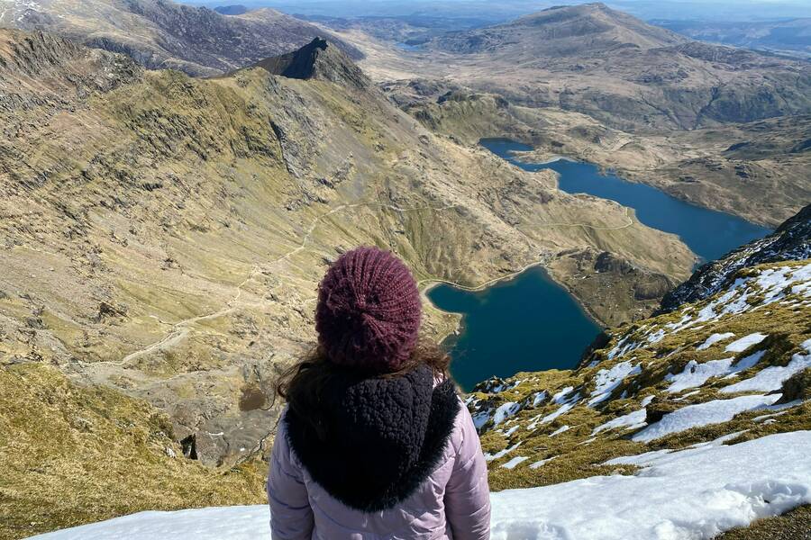 A person in a winter coat and knitted hat looks out over the dramatic mountain landscape of Yr Wyddfa above Llanberis, with rugged peaks, winding paths, and deep blue lakes below.