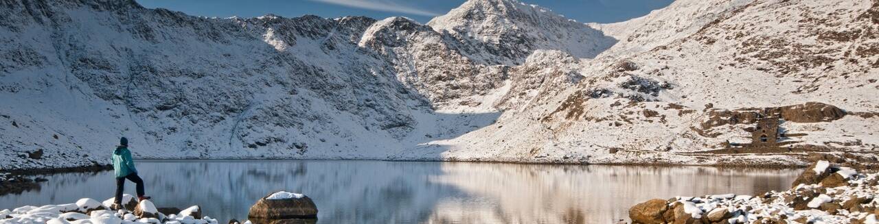 Hiker standing beside a snow-covered lake along the Llanberis Path on Snowdon, with the mountain peaks reflected in the still water.